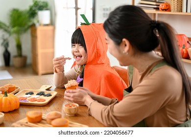 An Adorable Asian Girl In Halloween Costume Enjoys Eating Cupcake With Her Mom At Home. Halloween Activity Concept