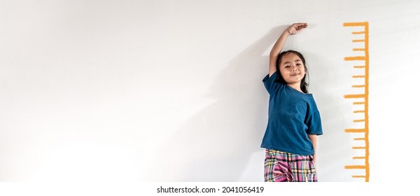 An Adorable Asian Little Child Girl Measuring The Height Growth With The Orange Drawing On The White Wall Background. Girl Power Future And Dream Concept Idea. Copy Space.