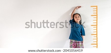 An adorable Asian little child girl measuring the height growth with the orange drawing on the white wall background. Girl power future and dream concept idea. Copy space.