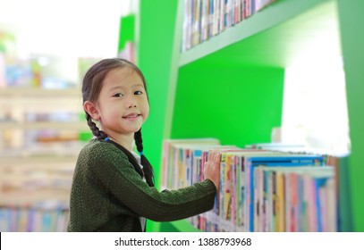 Adorable Asian Little Girl Looking For Book On Bookshelf At Library. Child Reaches For Books On A Shelf.