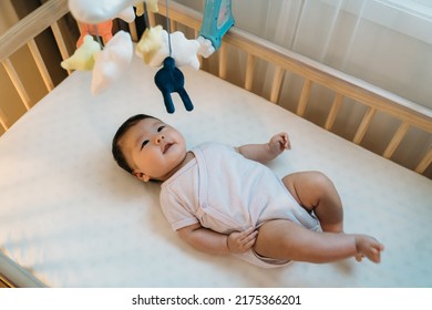 adorable asian newborn child is looking upward at the colorful hanging toys with curiosity while playing alone in the baby bed with white bedding at home. - Shutterstock ID 2175366201