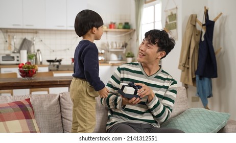 Adorable Asian Toddler Boy Covering His Dad’s Eyes And Showing Him A Gift From Behind On Father’s Day. The Father Opening The Box With Smile On Sofa