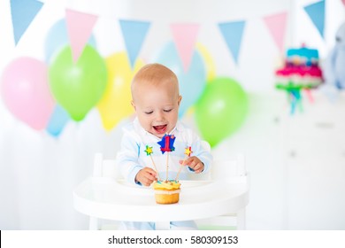 Adorable baby boy celebrating first birthday blowing candles on colorful cup cake. Kids birthday party decorated with balloons and pastel color banner. Child eating cake and candy