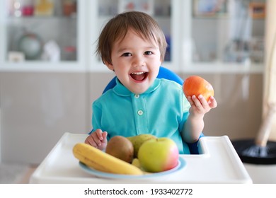 Adorable baby boy holding fresh orange in his hand with smiling face. Happy mixed race Asian-German child with healthy nutrition sitting on high chair at home.