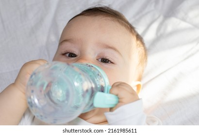 adorable baby boy laying on bed drinking water from plastic bottle with silicone head for mouth. toddler hold legs up. cute baby feet.cup with 2 handles,anti leak, straw wide mouth. 
