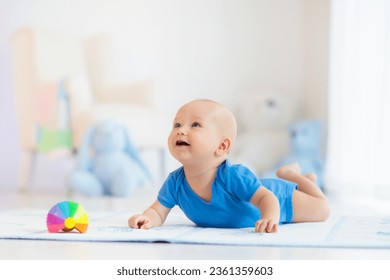 Adorable baby boy learning to crawl and playing with colorful rainbow ball toy in white sunny bedroom. Cute laughing child crawling on a play mat. Nursery, clothing and toys for little kids. 