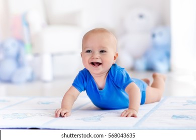Adorable baby boy learning to crawl and playing with colorful toy in white sunny bedroom. Cute laughing child crawling on a play mat. Nursery interior, clothing and toys for little kids 