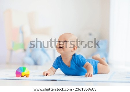 Adorable baby boy learning to crawl and playing with colorful rainbow ball toy in white sunny bedroom. Cute laughing child crawling on a play mat. Nursery, clothing and toys for little kids. 