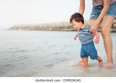 Adorable baby boy making his first steps on the beach by the seaside with his mother. One year old Infant learning how to walk on a summer vacation. Large copy space for text - Powered by Shutterstock