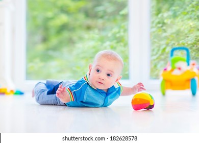 Adorable baby boy playing with a colorful ball and toy car in a sunny nursery with white furniture and white floor and a big garden view window