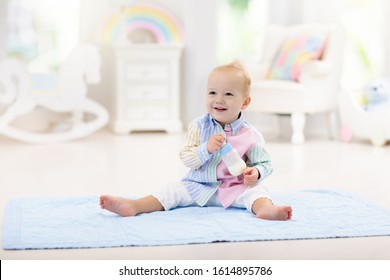 Adorable Baby Boy Playing On A Blue Floor Mat And Drinking Milk From A Bottle In A White Sunny Nursery With Rocking Chair And Bassinet. Bedroom Interior With Infant Crib. Formula Drink For Infant.