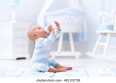 Adorable Baby Boy Playing On A Blue Floor Mat And Drinking Milk From A Bottle In A White Sunny Nursery With Rocking Chair And Bassinet. Bedroom Interior With Infant Crib. Formula Drink For Infant.