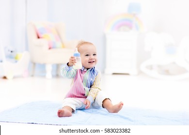 Adorable Baby Boy Playing On A Blue Floor Mat And Drinking Milk From A Bottle In A White Sunny Nursery With Rocking Chair And Bassinet. Bedroom Interior With Infant Crib. Formula Drink For Infant