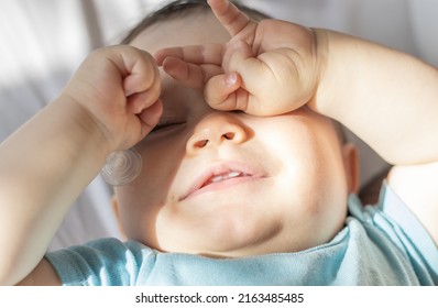 adorable baby boy rubs his eyes.sleepy cute baby boy with hand on eyes lying on bed.sunny rays on toddler pretty face.maternity. 2 teeths up.
