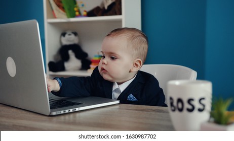 Adorable baby boy sitting by the table with the computer and boss’s mug, clapping his hands. Shelf with toys on the background. Baby boss concept. Having fun, successful, child’s portrait