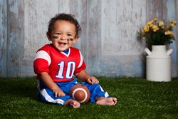 Adorable Baby Boy Sitting In The Grass, Wearing A Football Uniform.  