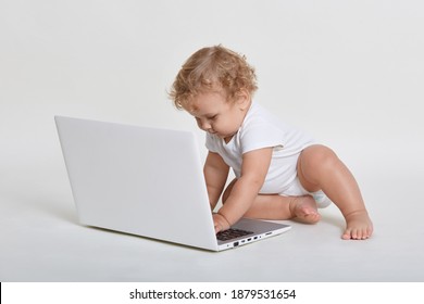 Adorable Baby Boy Sitting On Floor And Looking At Laptop Screen, Touching Its Keyboard, Kid Wearing White Bodysuit, Has Blond Wavy Hair, Tot Watching Cartoon Indoor.