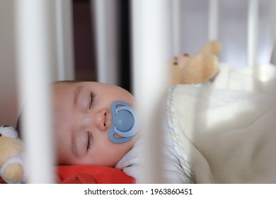 Adorable Baby Boy Sleeping With Pacifier On Mouth In Cot With Teddy Bear Toy. Mixed Race Asian-German Infant Lying In Crib. Little Child Sleep With Blanket.