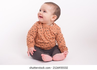 Adorable baby boy wearing animal print sweater and grey pants sitting on white background looking aside and smiling to his mom. Baby looking upwards.