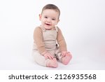 Adorable baby boy wearing beige overalls sitting on white background looking at camera and smiling. 