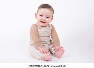 Adorable bebé con overoles beige sobre fondo blanco mirando a la cámara y sonriendo. 