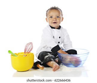 An adorable baby boy wearing a chef's jacket and black pants, playing with apples and cooking utensils, and messed up with white flour.  On a white background.