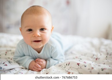 Adorable Baby Boy In White Sunny Bedroom In Winter Morning. Newborn Child Relaxing In Bed. Family Morning At Home. Newborn Kid During Tummy Time Smiling Happily At Home