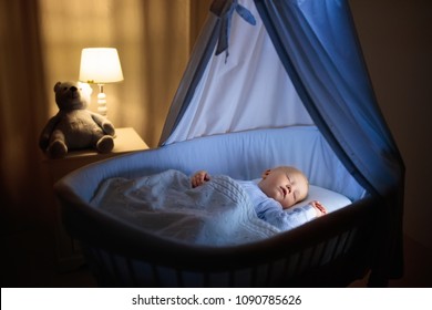 Adorable Baby Drinking Milk In Blue Bassinet With Canopy At Night. Little Boy In Pajamas With Formula Bottle Getting Ready To Sleep In Dark Room With Crib, Lamp And Toy Bear. Bed Time Drink For Kids.