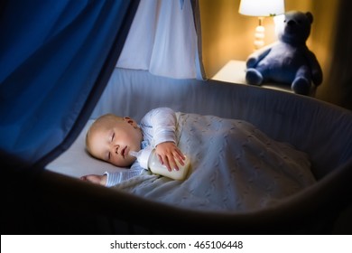 Adorable Baby Drinking Milk In Blue Bassinet With Canopy At Night. Little Boy In Pajamas With Formula Bottle Getting Ready To Sleep In Dark Room With Crib, Lamp And Toy Bear. Bed Time Drink For Kids.