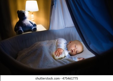 Adorable Baby Drinking Milk In Blue Bassinet With Canopy At Night. Little Boy In Pajamas With Formula Bottle Getting Ready To Sleep In Dark Room With Crib, Lamp And Toy Bear. Bed Time Drink For Kids.