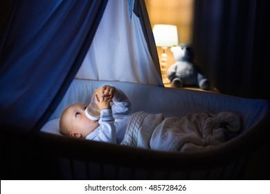 Adorable Baby Drinking Milk In Blue Bassinet With Canopy At Night. Little Boy In Pajamas With Formula Bottle Getting Ready To Sleep In Dark Room With Crib, Lamp And Toy Bear. Bed Time Drink For Kids.