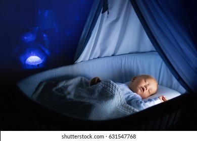 Adorable Baby Drinking Milk In Blue Bassinet With Canopy At Night. Little Boy In Pajamas With Formula Bottle Getting Ready To Sleep In Dark Room With Crib, Lamp And Toy Bear. Bed Time Drink For Kids.