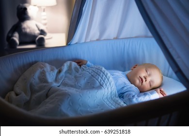 Adorable Baby Drinking Milk In Blue Bassinet With Canopy At Night. Little Boy In Pajamas With Formula Bottle Getting Ready To Sleep In Dark Room With Crib, Lamp And Toy Bear. Bed Time Drink For Kids.