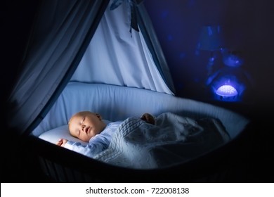 Adorable Baby Drinking Milk In Blue Bassinet With Canopy At Night. Little Boy In Pajamas With Formula Bottle Getting Ready To Sleep In Dark Room With Crib, Lamp And Toy Bear. Bed Time Drink For Kids.