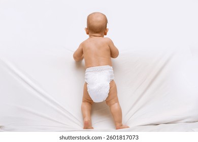 Adorable bebé en un pañal volante de pie en una cama blanca suave. Desarrollo temprano e hito en la infancia. Vista trasera.  El bebé aprende a pararse, desarrollando equilibrio y fuerza. 