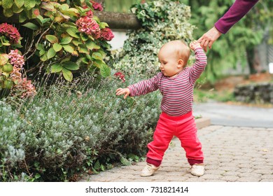 Adorable Baby Girl Of 9-12 Months Old Playing Outside, Wearing Pink Body And Joggers, Holding Mother's Hand. Child's First Steps, Kid Learning To Walk