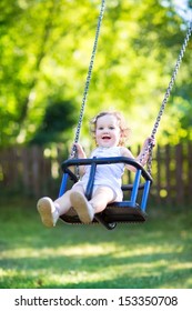 Adorable baby girl with big beautiful eyes and curly hair having fun on a swing ride at a playground in a sunny summer park