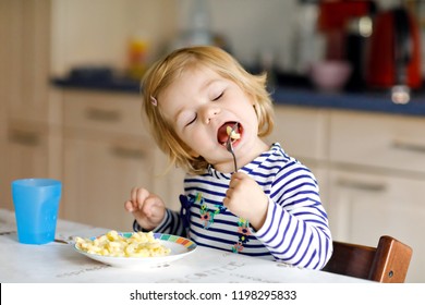 Adorable Baby Girl Eating From Spoon Noodle, Pasta Macaroni. Cute Healthy Toddler Child, Daughter With Spoon Sitting In Highchair And Learning To Eat By Itself In Domestic Kitchen Or Nursery