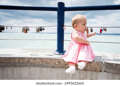 Adorable baby girl in pink dress sitting by the seaside. Seaside view with padlocks on railings. Cute moment captured outdoors. - Powered by Shutterstock