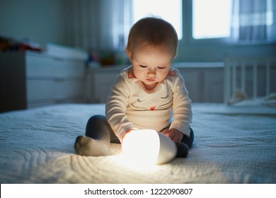 Adorable Baby Girl Playing With Bedside Lamp In Nursery. Happy Kid Sitting On Bed With Nightlight. Little Child At Home In The Evening Before Sleep