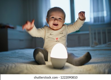 Adorable Baby Girl Playing With Bedside Lamp In Nursery. Happy Kid Sitting On Bed With Nightlight. Little Child At Home In The Evening Before Sleep