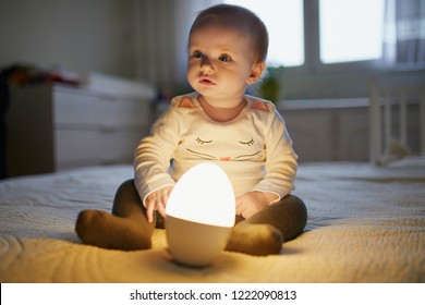 Adorable baby girl playing with bedside lamp in nursery. Happy kid sitting on bed with nightlight. Little child at home in the evening before sleep