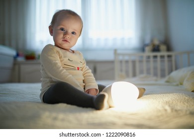 Adorable Baby Girl Playing With Bedside Lamp In Nursery. Happy Kid Sitting On Bed With Nightlight. Little Child At Home In The Evening Before Sleep