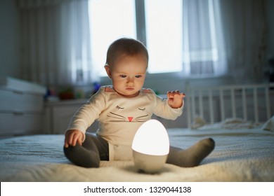 Adorable Baby Girl Playing With Bedside Lamp In Nursery. Happy Kid Sitting On Bed With Nightlight. Little Child At Home In The Evening Before Sleep