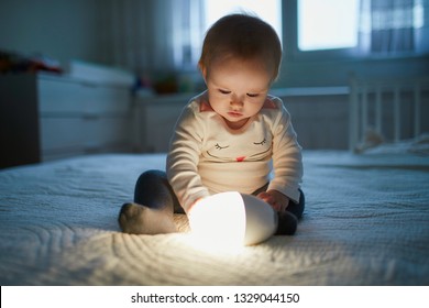 Adorable Baby Girl Playing With Bedside Lamp In Nursery. Happy Kid Sitting On Bed With Nightlight. Little Child At Home In The Evening Before Sleep