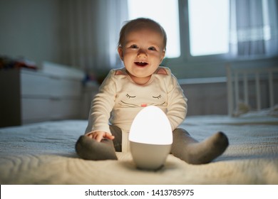 Adorable Baby Girl Playing With Bedside Lamp In Nursery. Happy Kid Sitting On Bed With Nightlight. Little Child At Home In The Evening Before Sleep