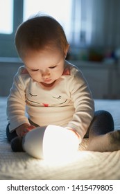 Adorable Baby Girl Playing With Bedside Lamp In Nursery. Happy Kid Sitting On Bed With Nightlight. Little Child At Home In The Evening Before Sleep