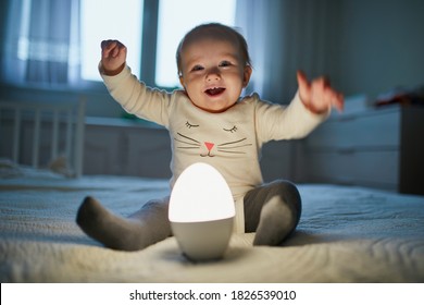 Adorable Baby Girl Playing With Bedside Lamp In Nursery. Happy Kid Sitting On Bed With Nightlight. Little Child At Home In The Evening Before Sleep