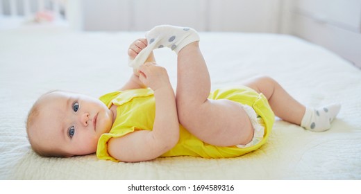 Adorable Baby Girl Removing Sock From Her Foot. Little Child Having Fun. Infant Kid In Sunny Nursery