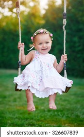 Adorable baby girl wearing a white dress enjoying a swing ride on a playground in a park on a nice sunny summer day